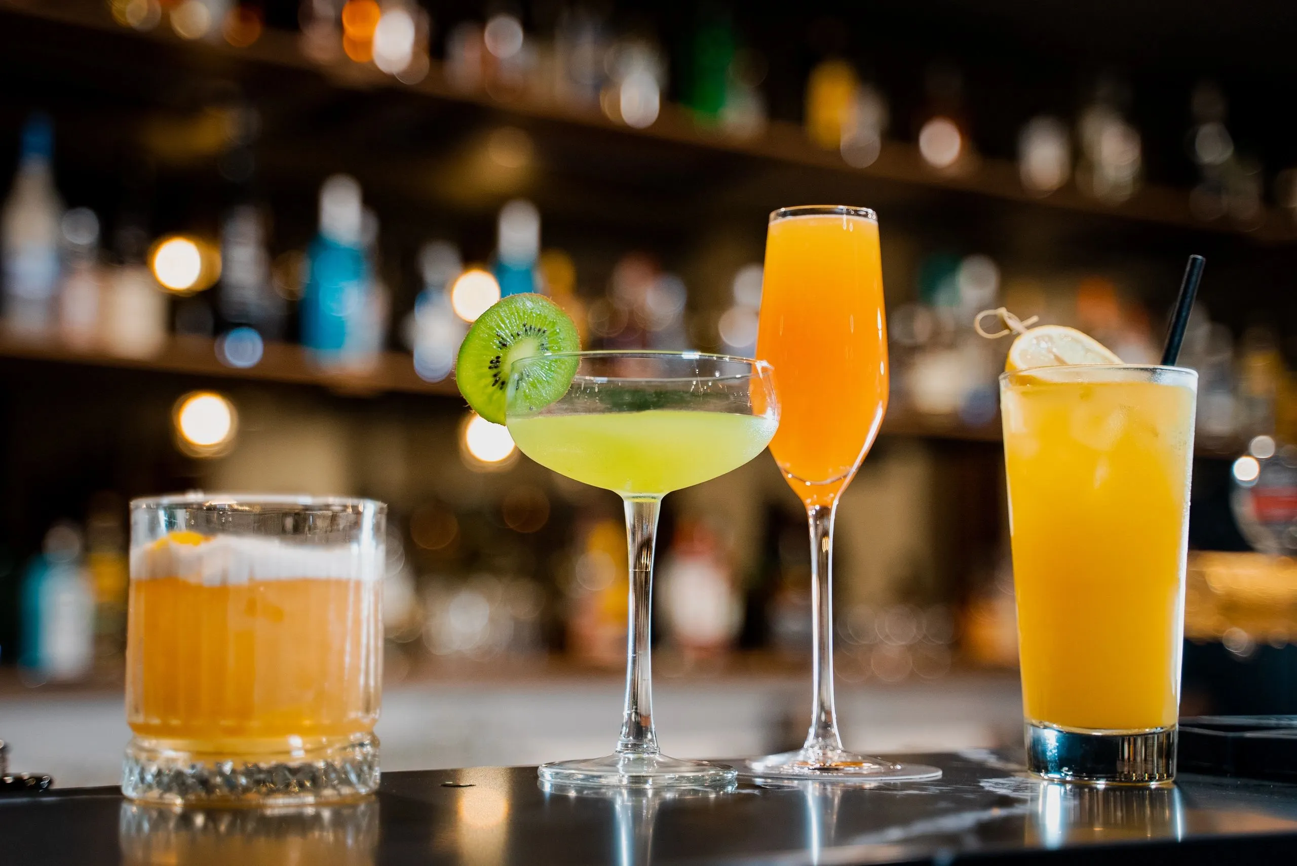 Close up of a cocktail in a coupe with a slice of kiwi, the back bar glowing behind