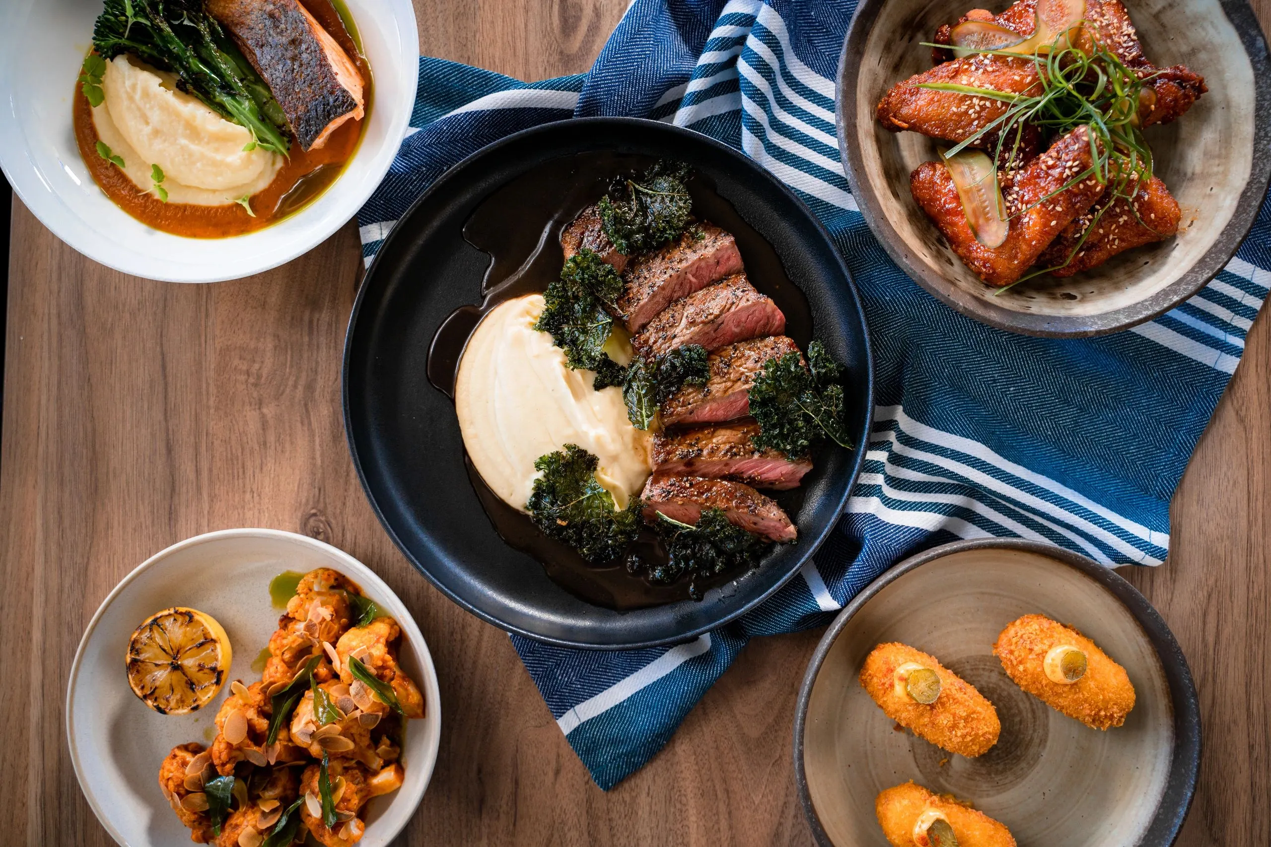 A table of T-bone dishes from above: steak with mash and kale, salmon, glazed wings, cauliflower and croquettes