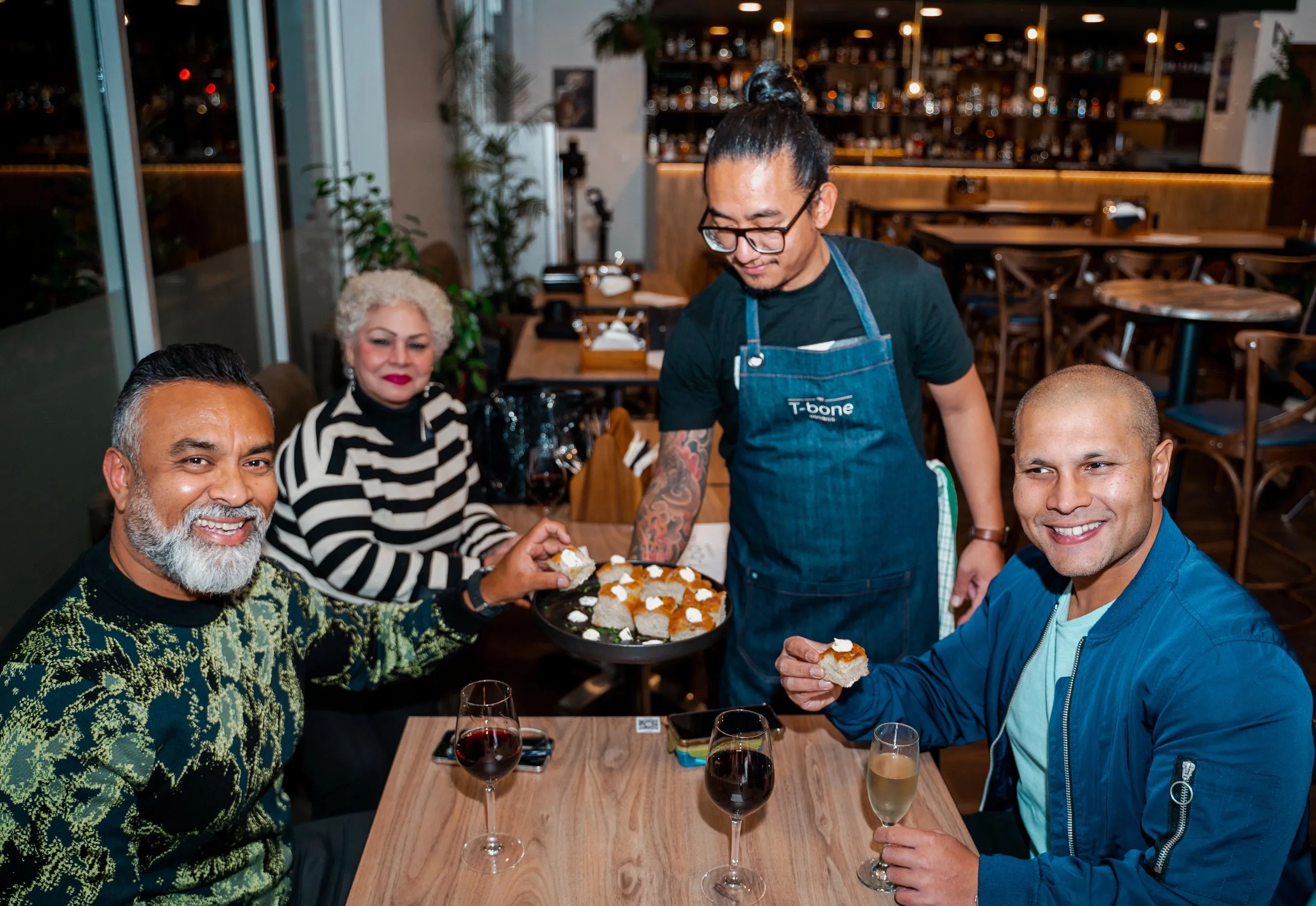A T-bone waiter in a denim apron serving bread to smiling guests at a table
